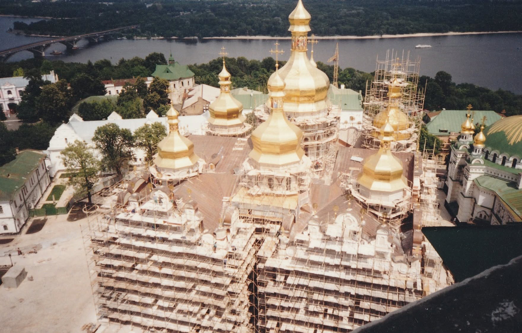 Colour aerial photograph of the Dormition Cathedral being rebuilt: gold domes installed, lower walls in scaffolding, the Dnipro behind.