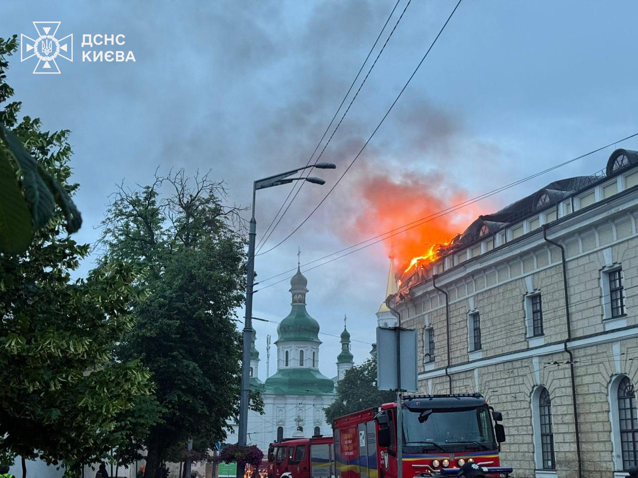 Firefighters and engines on the street below the burning roof of the Mystetskyi Arsenal at dawn, the green domes of a Lavra church behind, 15 June 2026.