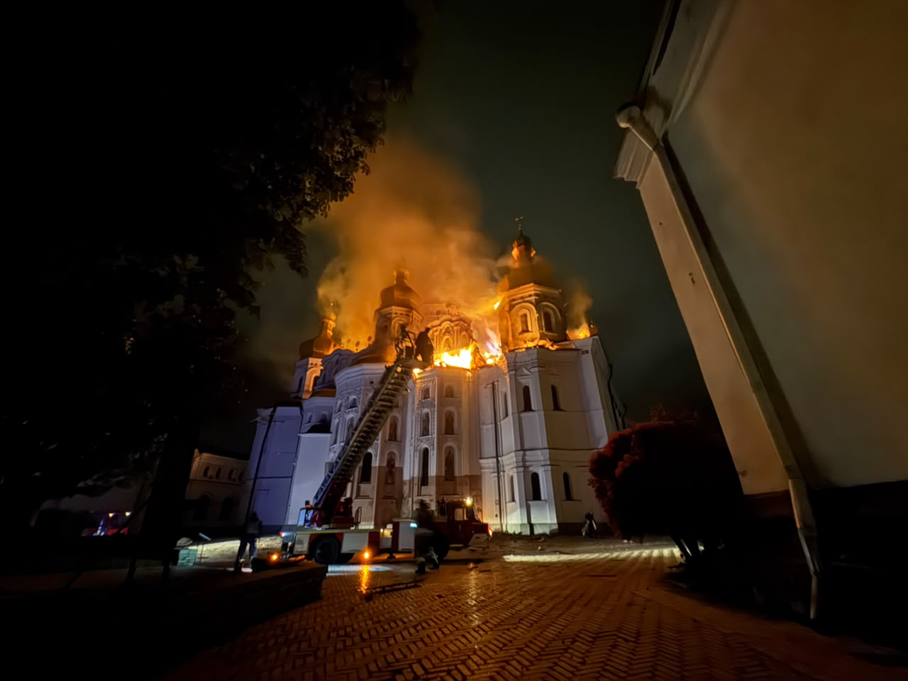 The Dormition Cathedral ablaze at night, large flames erupting from the roof between the domes as an aerial ladder and fire engine work below, 15 June 2026.