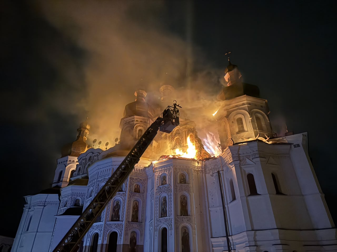 An aerial ladder reaching the burning roof of the Dormition Cathedral at night, flames among the frescoed apse and gilded domes, 15 June 2026.