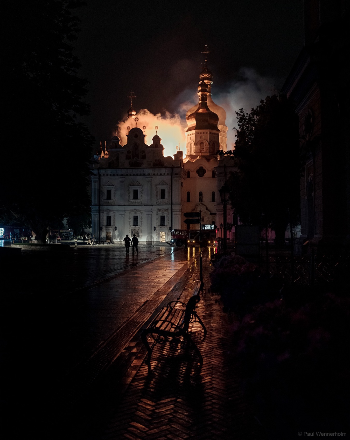 A wet path leading toward the Dormition Cathedral at night, fire glowing behind its domes through the smoke, a bench in silhouette and firefighters near the floodlit building, 15 June 2026.