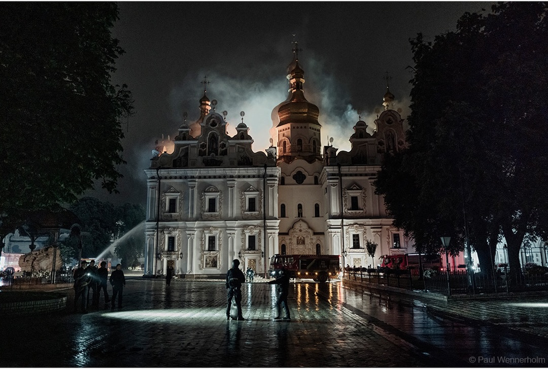 The floodlit facade of the Dormition Cathedral at night with fire glowing behind the domes and firefighters hosing the building across a wet square, 15 June 2026.
