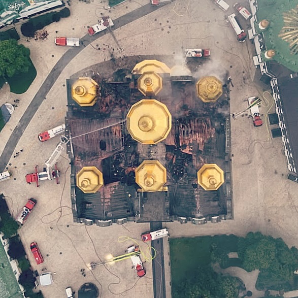 Top-down drone view of the Dormition Cathedral the day after the fire: the seven gilded domes intact while the roof between them is burned away to charred timbers, fire engines ringing the building, 15 June 2026.