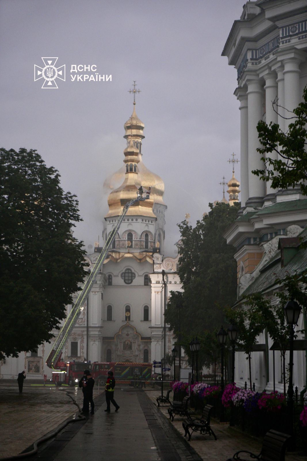 A fire-service aerial ladder reaching the gilded central dome of the Dormition Cathedral as a firefighter douses smoke, dawn of 15 June 2026.
