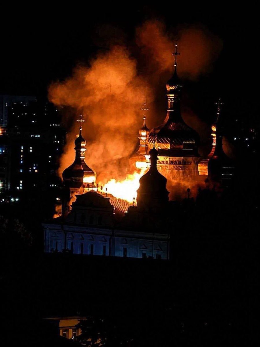 The gilded domes and crosses of the Kyiv-Pechersk Lavra in silhouette against rooftop flames and heavy smoke, a lit residential tower behind, telephoto night view of 15 June 2026.