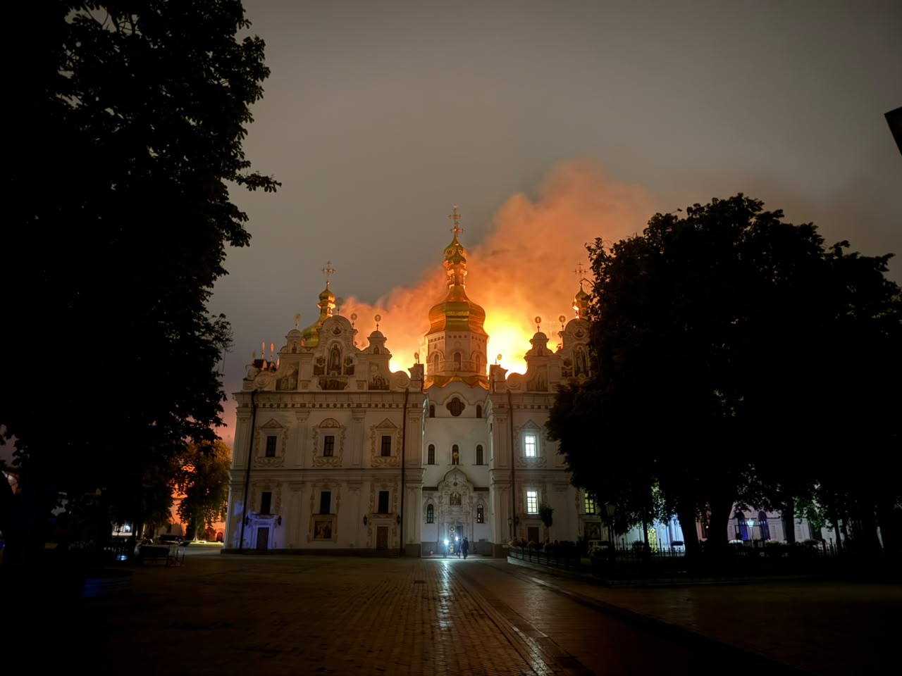 The Dormition Cathedral of the Kyiv-Pechersk Lavra at night, flames and smoke rising behind its gilded domes, framed by dark trees, after the Russian drone strike of 15 June 2026.