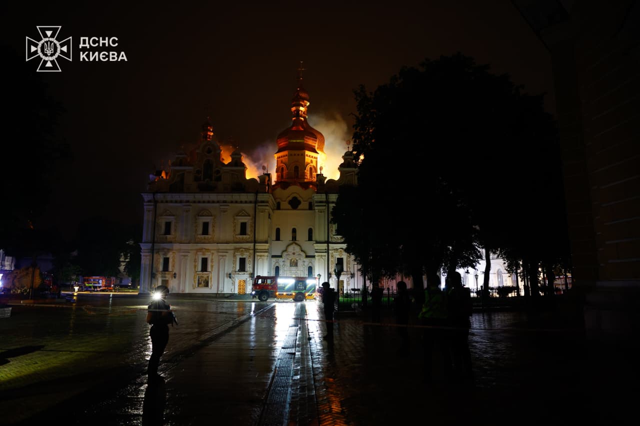 The illuminated baroque facade of the Dormition Cathedral with fire glowing behind it and a fire engine on the wet square, night of 15 June 2026.