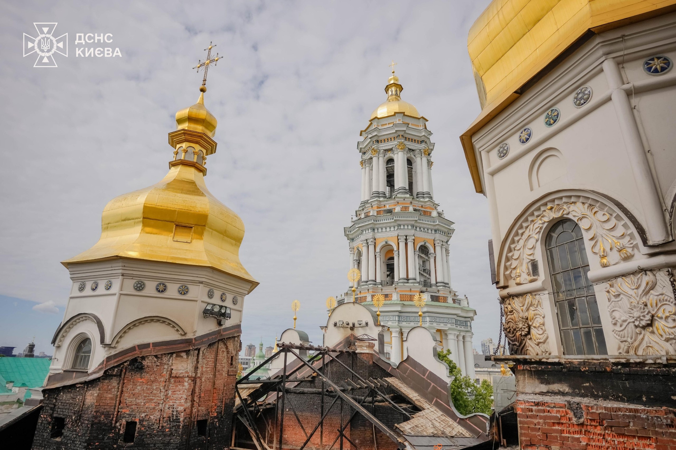 Daytime view from roof level of the Dormition Cathedral showing charred rafters and exposed brick, the Great Bell Tower and gilded domes beyond, the day after the strike, 16 June 2026.