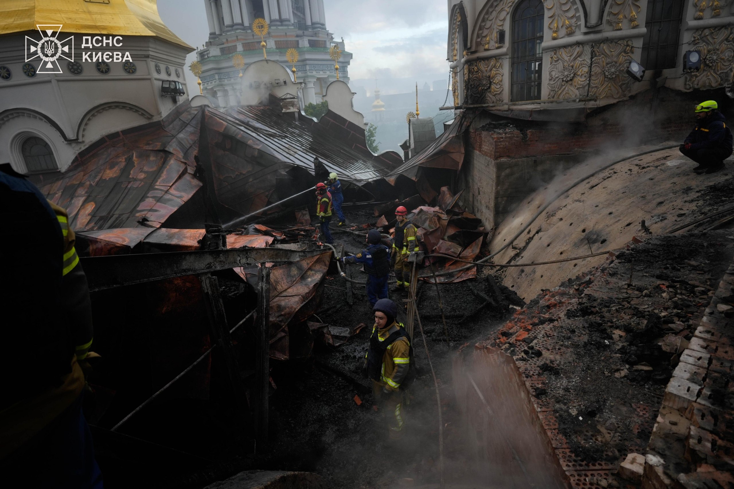 Firefighters working amid charred, collapsed roof timbers on the Dormition Cathedral, its gilded domes and white walls above, 15 June 2026.